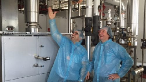 Due persone con abbigliamento protettivo si trovano in un ambiente industriale e osservano un impianto con tubazioni e componenti tecnici.