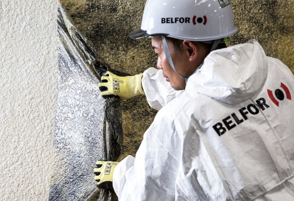 A man in a white jacket and safety glasses peels a film from a wall to remove soot after a fire.