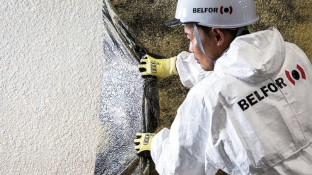 A man in a white jacket and safety glasses peels a film from a wall to remove soot after a fire.