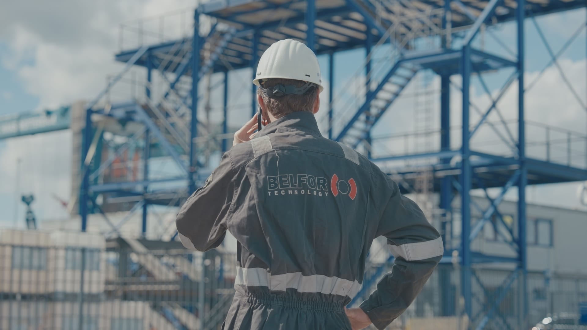 Een medewerker van BELFOR Technology met een helm en een beschermend pak, met een telefoon in zijn hand, staande voor een bedrijfsgebouw