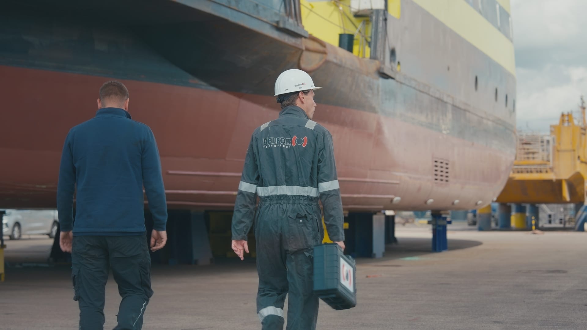 Een technicus van BELFOR, gekleed in helm en overall, en een collega lopen langs een groot schip in een droogdok